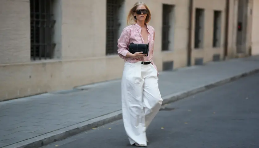 Kobieta w stylowym, różowo-białym paszku Massimo Dutti, szerokich białych spodniach i okularach przeciwsłonecznych idzie ulicą z czarną kopertówką.