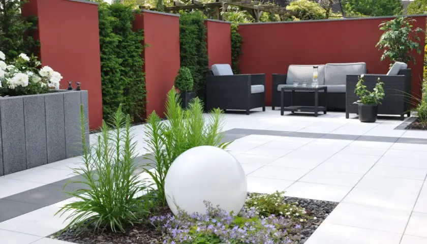 Moderner Garten ohne Rasen mit roten Mauern, Sitzgruppe und weißer Kugel im Blumenbeet.