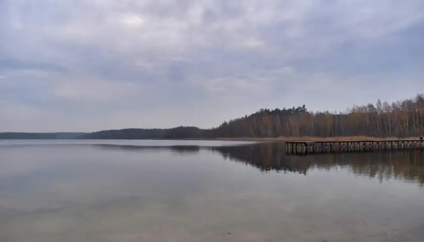 Spokojne jezioro Kamionki odbija pochmurne niebo i lasy. Drewniane molo wchodzi w głąb wody.