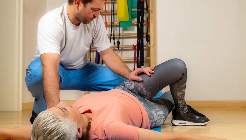 Fizjoterapeuta pomaga pacjentce wykonać test Patricka. Kobieta leży na macie z ugiętymi nogami, a mężczyzna delikatnie naciska na jej kolano.