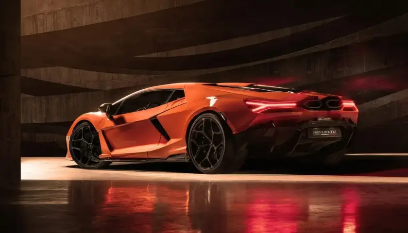Pomarańczowy Lamborghini Revuelto z czarnymi felgami, stojący w ciemnym, betonowym garażu, z czerwonymi światłami odbijającymi się od podłogi.