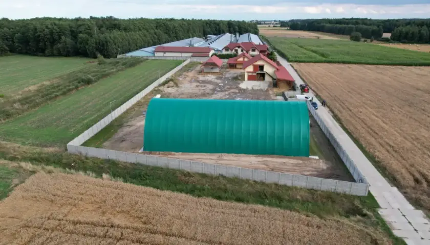 Zielona hala namiotowa na terenie gospodarstwa. Obok budynki mieszkalne i gospodarcze, otoczone polami uprawnymi. Grunty rolne zabudowane.