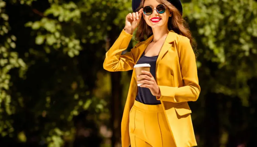 Une femme en costume jaune vif, chapeau et lunettes de soleil, tenant un café. Parfait pour savoir comment s'habiller quand il fait 14 degrés.