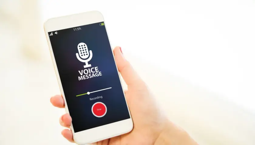 Dłoń trzyma smartfon z włączonym dyktafonem w telefonie. Na ekranie widnieje ikona mikrofonu i napis "VOICE MESSAGE".
