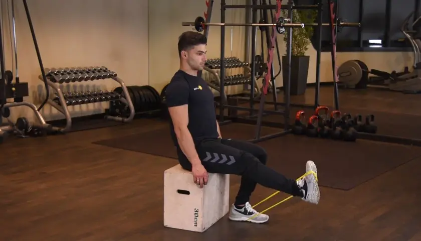 Mężczyzna wykonuje ćwiczenie sissy squat z gumą oporową na nogach, siedząc na skrzyni w siłowni.