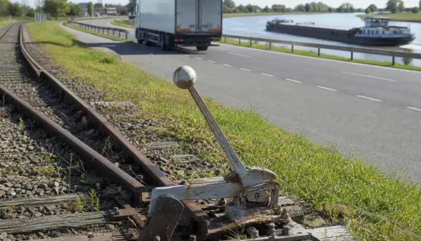Alte Weiche neben Gleisen. Ein LKW und ein Frachtschiff sind auf der Straße und dem Wasser zu sehen, verschiedene Verkehrsmittel.