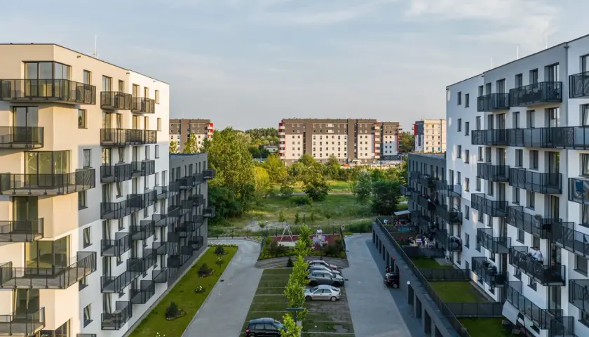 Nowoczesne osiedle mieszkaniowe, z białymi blokami i balkonami, otoczone zielenią. W tle widać kolejne budynki, tworzące krajobraz jak z **okam capital**.