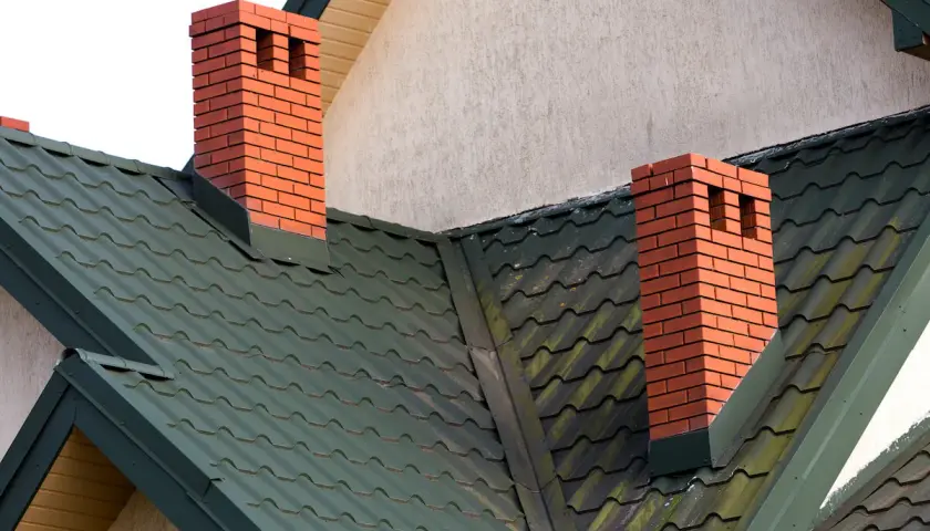 Dwa ceglane kominy na zielonym dachu. Warto sprawdzić odległość komina od granicy działki przed budową.