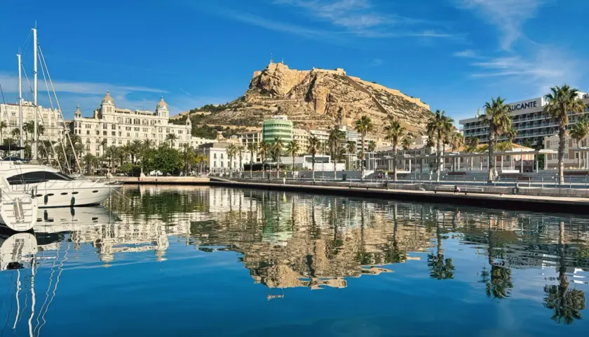 Alicante, luty. Słoneczny dzień nad zatoką z odbiciem zamku Santa Barbara i palm w krystalicznie czystej wodzie.