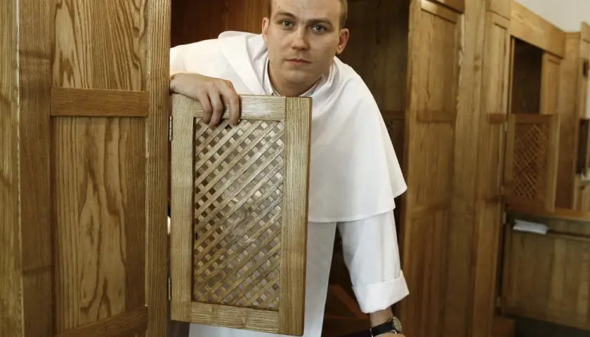 Młody zakonnik w białej sutannie otwiera kratkę konfesjonału, gotów wysłuchać spowiedzi.