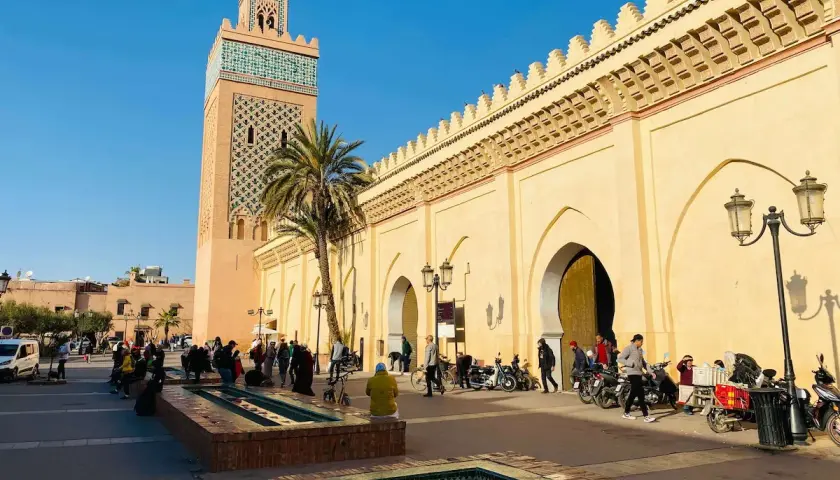 La moschea Koutoubia domina la piazza, con la sua torre decorata e le arcate del souk di Marrakech. Gente e motorini animano la scena.