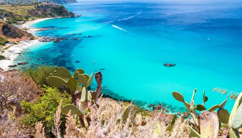 A stunning view of Reggio Calabria beaches, with turquoise waters, white sand, and lush green cliffs dotted with cacti.