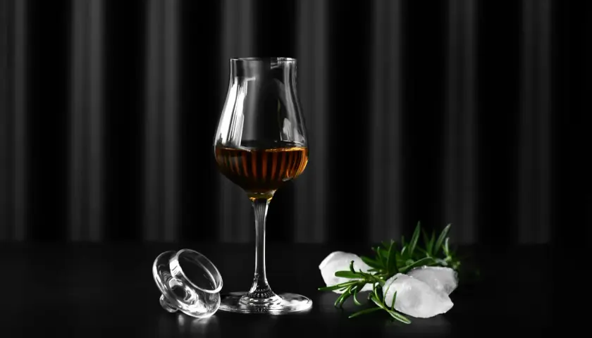 Szklanka Glencairn z bursztynowym trunkiem, obok leży jej zakrętka, lód i rozmaryn. Jak dobrać odpowiednie szkło Glencairn vs tumbler vs copita.