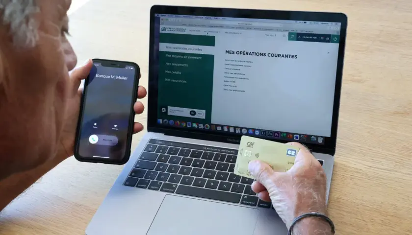 Un homme tient un téléphone et une carte bancaire, devant un ordinateur affichant le site du Crédit Agricole. Il cherche peut-on annuler un paiement par carte bancaire sur internet.
