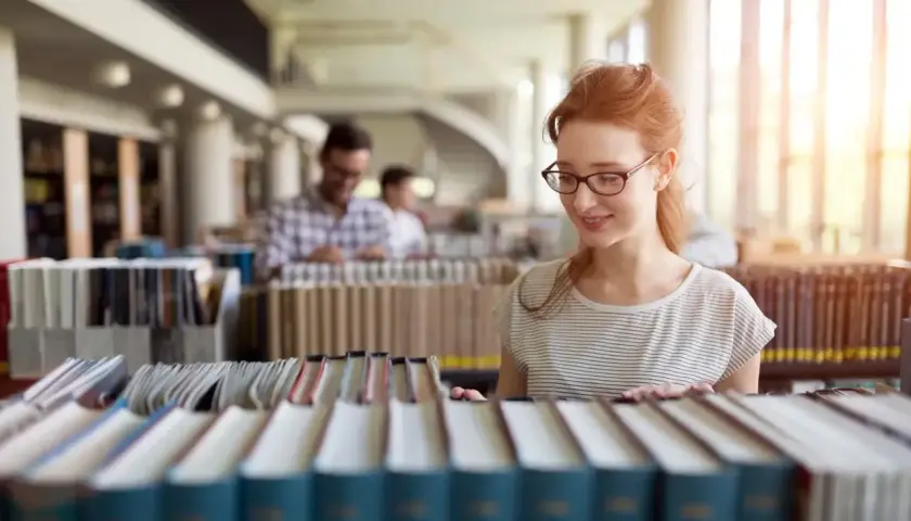 Studentka z rudymi włosami w okularach przegląda książki w bibliotece, szukając materiałów do prawo studia.