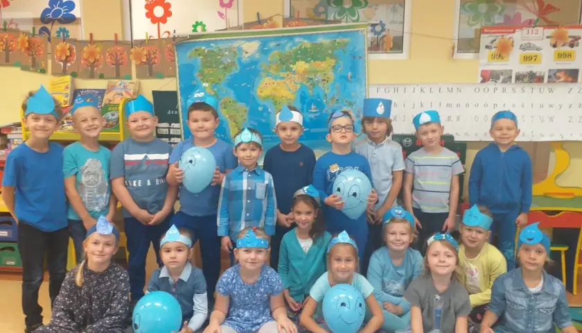 Grupa przedszkolaków w niebieskich czapeczkach z kropelkami wody, trzymających balony.
