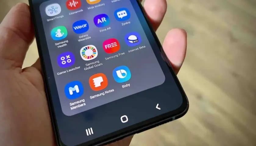Dłoń trzyma smartfon z widocznym folderem aplikacji, w tym ikoną Bixby.