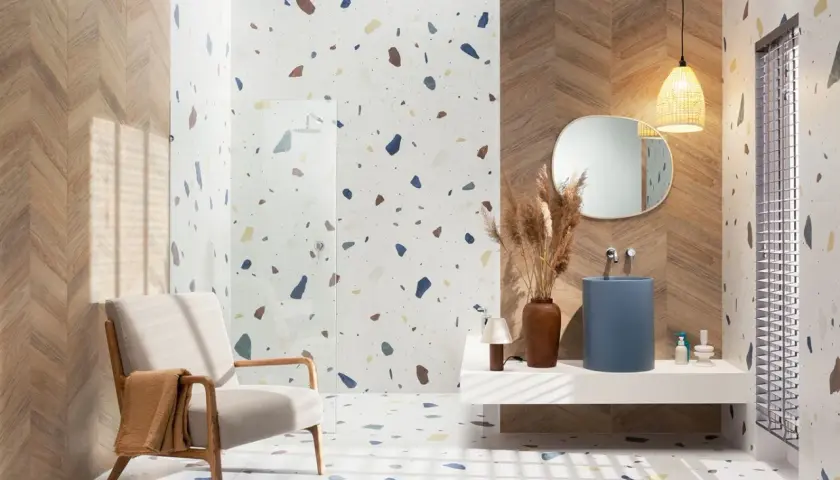 Nowoczesna łazienka z podłogą i ścianami w stylu **terrazzo**, drewnianymi akcentami, fotelem i niebieską umywalką.