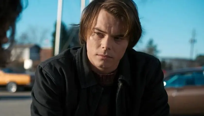 Charlie Heaton w ciemnej kurtce i swetrze, zamyślony, na tle błękitnego nieba i samochodów.