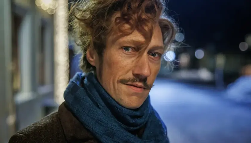 Man med rufsigt hår och mustasch, klädd i brun rock och blå halsduk. Kanske en del av rollistan i jakten på en hemlighet i vintermörkret.