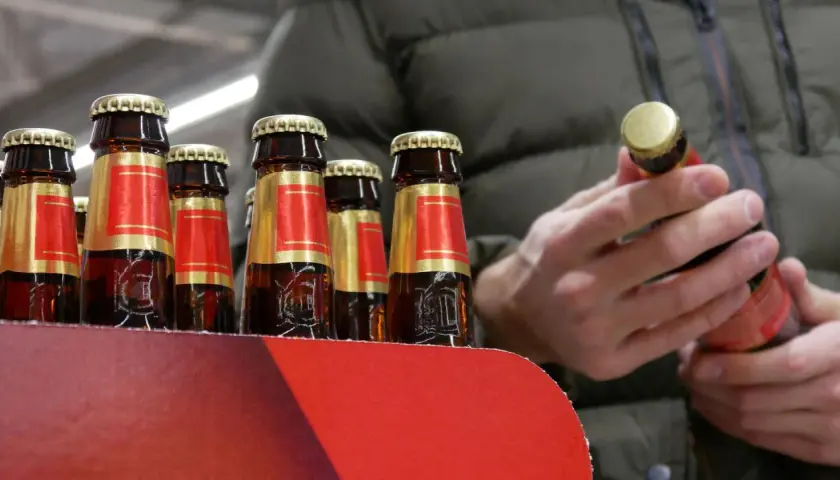 Rząd butelek piwa w czerwono-złotych etykietach. Człowiek w zielonej kurtce trzyma jedną butelkę, zastanawiając się, ile kosztuje jedno piwo.