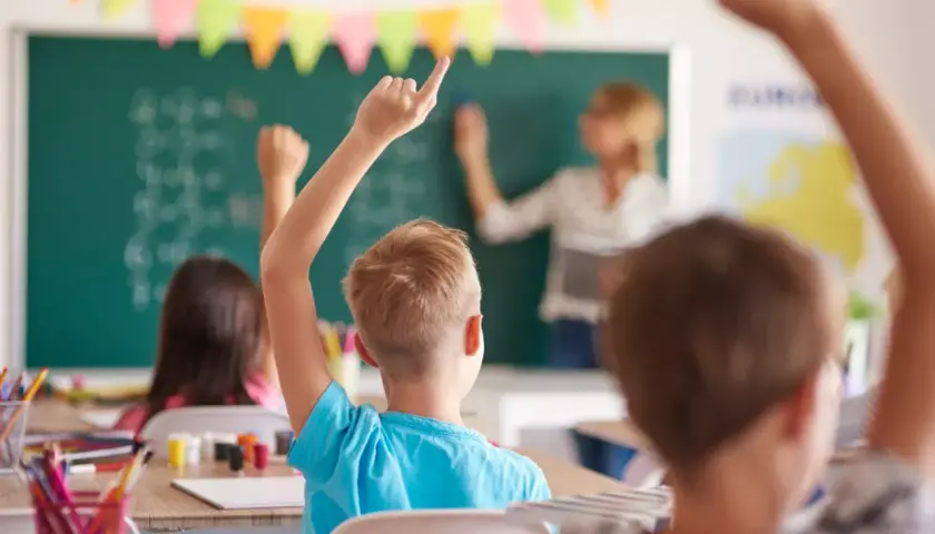 Uczniowie z podniesionymi rękami słuchają uważnie, co mówi wychowawca przy tablicy.