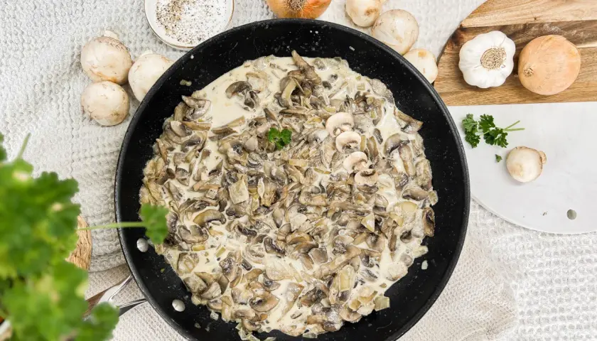 Kremowy sos pieczarkowy na patelni, idealny do makaronu, ziemniaków czy mięsa. Obok składniki: pieczarki, cebula, czosnek, natka pietruszki.