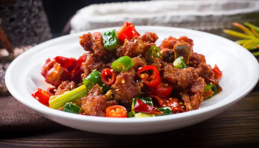 Chrupiący kurczak po kantońsku z papryczkami chili i zieloną cebulką na białym talerzu.