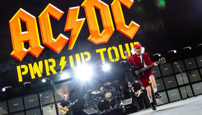 AC/DC na koncercie w Polsce. Wokalista w czerwonym stroju skacze z gitarą na tle gigantycznego logo zespołu i ściany wzmacniaczy.