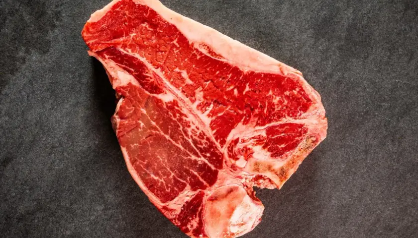 Una succulenta bistecca di manzo T-bone, con la sua marmorizzazione di grasso, pronta per essere cucinata su una superficie scura.