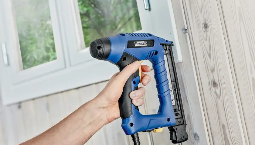 Hand håller en blå kompressor spikpistol mot en trävägg.