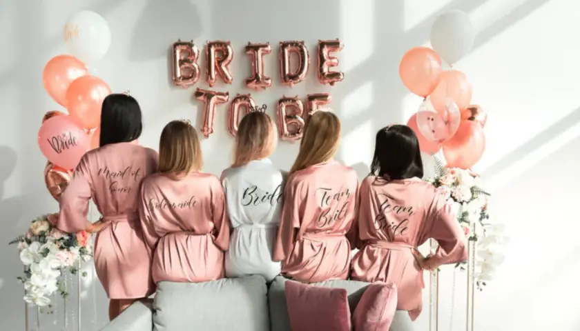 Pięć kobiet w satynowych szatkach, idealny prezent na wieczór panieński z klasą. Balony i napisy "Bride to Be" tworzą świąteczną atmosferę.