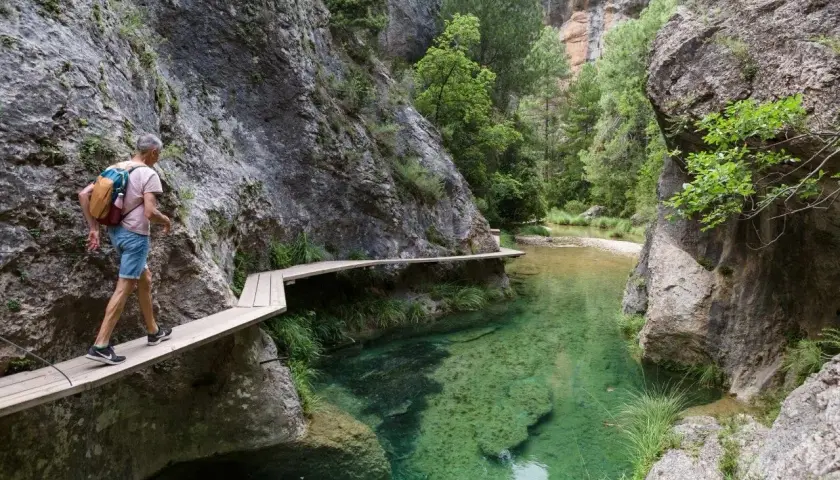 Hombre camina por pasarela de madera en la ruta parrizal de beceite, junto a un río de aguas cristalinas y paredes rocosas.