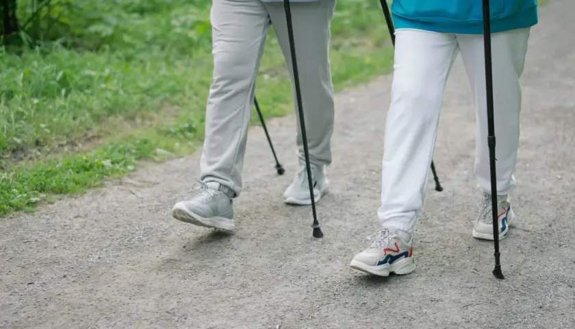 Dwie osoby uprawiające **nordic walking** na żwirowej ścieżce, otoczone zielenią.