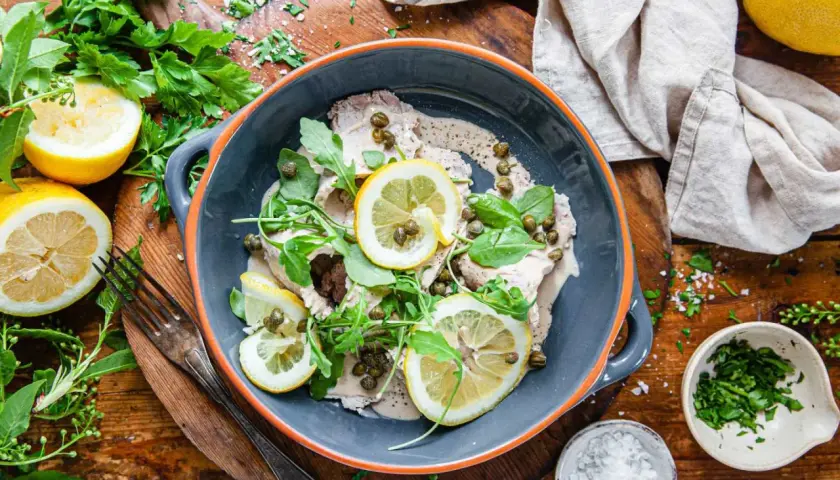 Vitello tonnato i en grå skål, garnerad med citronskivor, kapris och ruccola. Serveras med färska örter och citroner på ett träbord.