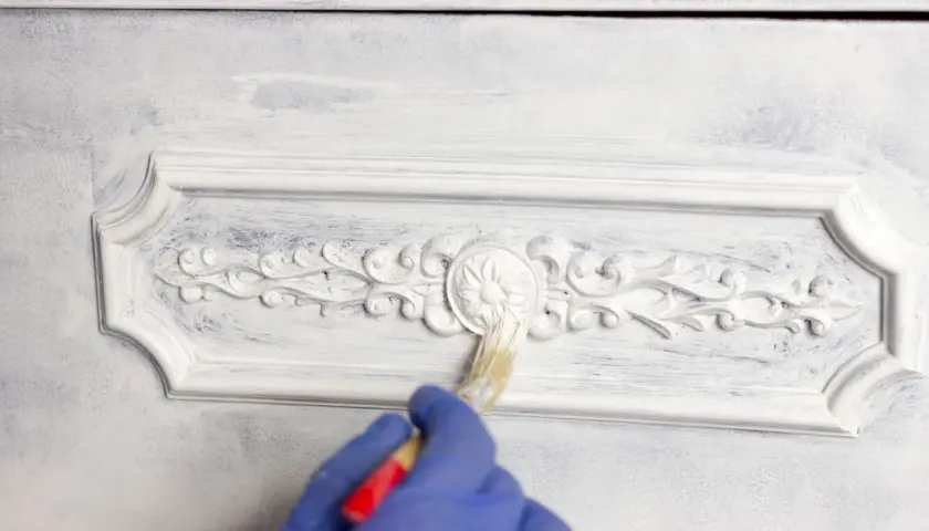 Mano con guanto blu che applica chalk paint fai da te su un mobile con decorazioni in rilievo, creando un effetto shabby chic.