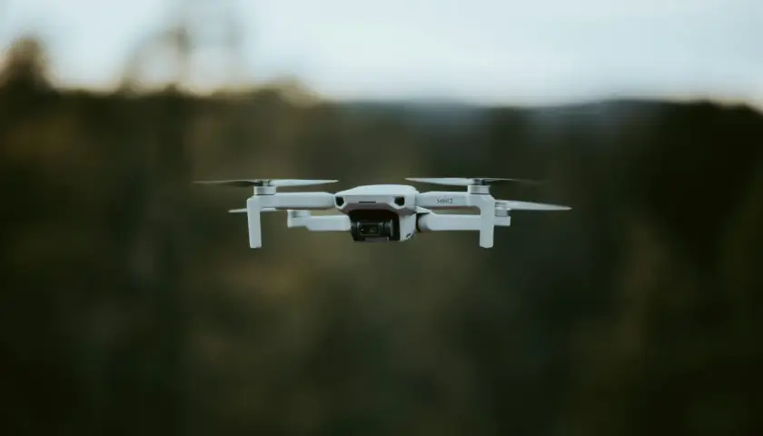 Biały dron DJI Mini 2 w locie, z widocznym obiektywem kamery. Tło to rozmyty las i niebo.