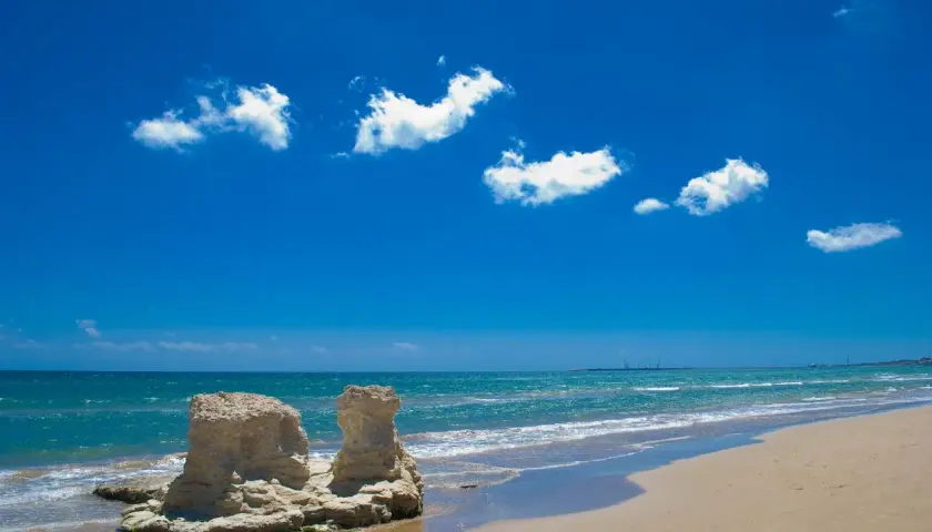 Spiagge attrezzate Sicilia: rocce scolpite dal mare e sabbia dorata sotto un cielo azzurro con nuvole bianche.