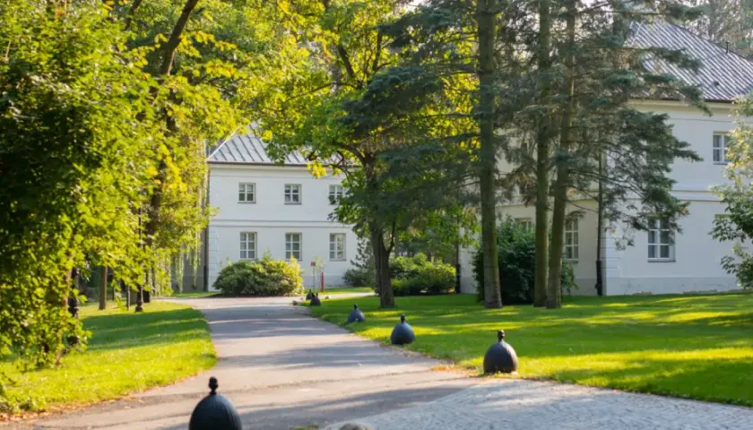 Biały dworek otoczony zielenią, z alejką i ozdobnymi kulami. Miejsce, gdzie nagrywano "Nad Niemnem".