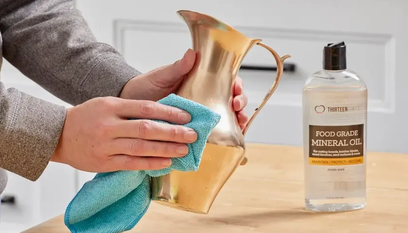Hands polish a brass pitcher with a blue cloth, showing how to clean brass with mineral oil.