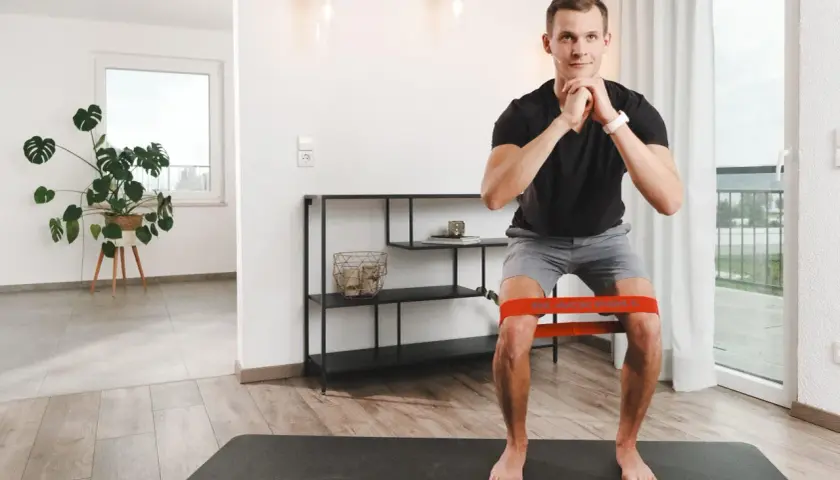 Mann macht Kniebeugen mit Widerstandsband. Die tiefe Flexion im Knie wird durch das Band unterstützt und erschwert.