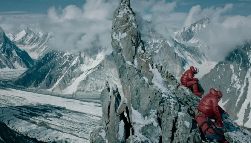 Dwóch alpinistów w czerwonych kurtkach odpoczywa na skalistej grani, podziwiając lodowiec i ośnieżone szczyty. Widok na berbeka broad peak zapiera dech w piersiach.