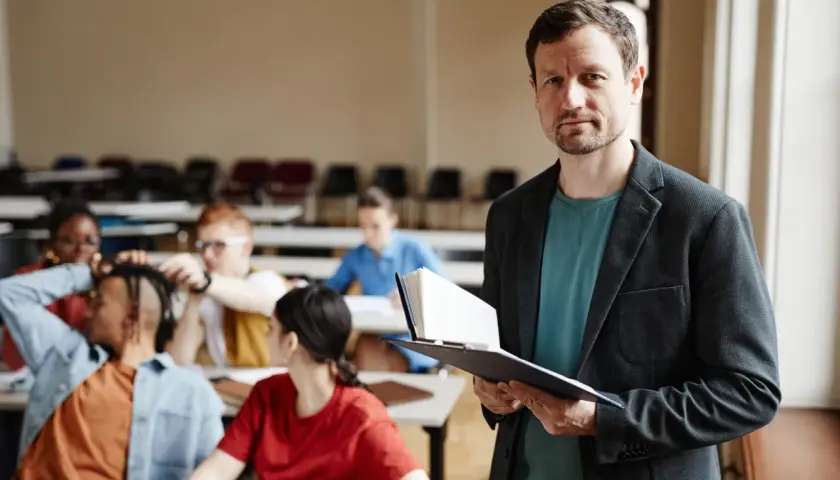 Nauczyciel z książką w ręku, przygotowujący się do lekcji. W tle widać uczniów, co sugeruje jego pensum nauczyciela.