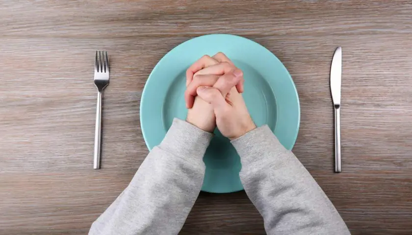 Manos entrelazadas sobre plato vacío, tenedor y cuchillo a los lados. Quizás piensa que no cenar adelgaza.