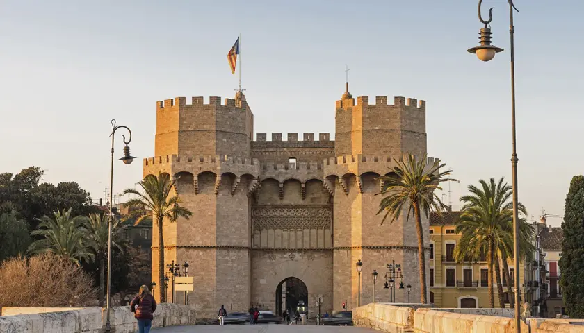 Las **Torres de Serranos**, imponentes puertas de la antigua muralla de Valencia, se alzan majestuosas bajo un cielo despejado. Palmeras y farolas flanquean el camino.