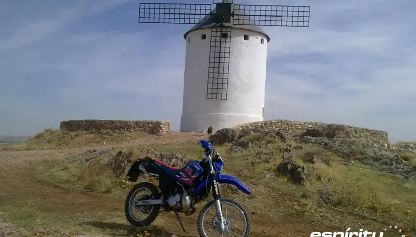 Yamaha DT 125 RE azul y roja aparcada junto a un molino de viento blanco en un paisaje rocoso.