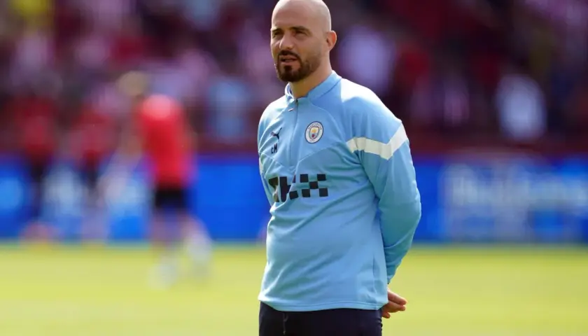Łysy mężczyzna z brodą, trener Manchester City, w błękitnej bluzie treningowej, stoi na boisku.