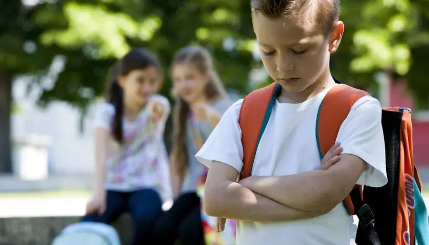 Ein Junge mit Rucksack steht traurig da, während zwei Mädchen ihn im Hintergrund auslachen. Ein trauriges Bild von Mobbing in der Grundschule.