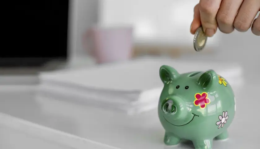Ręka wrzuca monetę do zielonej świnki skarbonki. To symbol oszczędzania i finansowego zabezpieczenia.