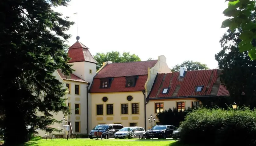 Pałac w Krokowej z czerwoną wieżą i dachami, otoczony zielenią. Przed budynkiem zaparkowane samochody.